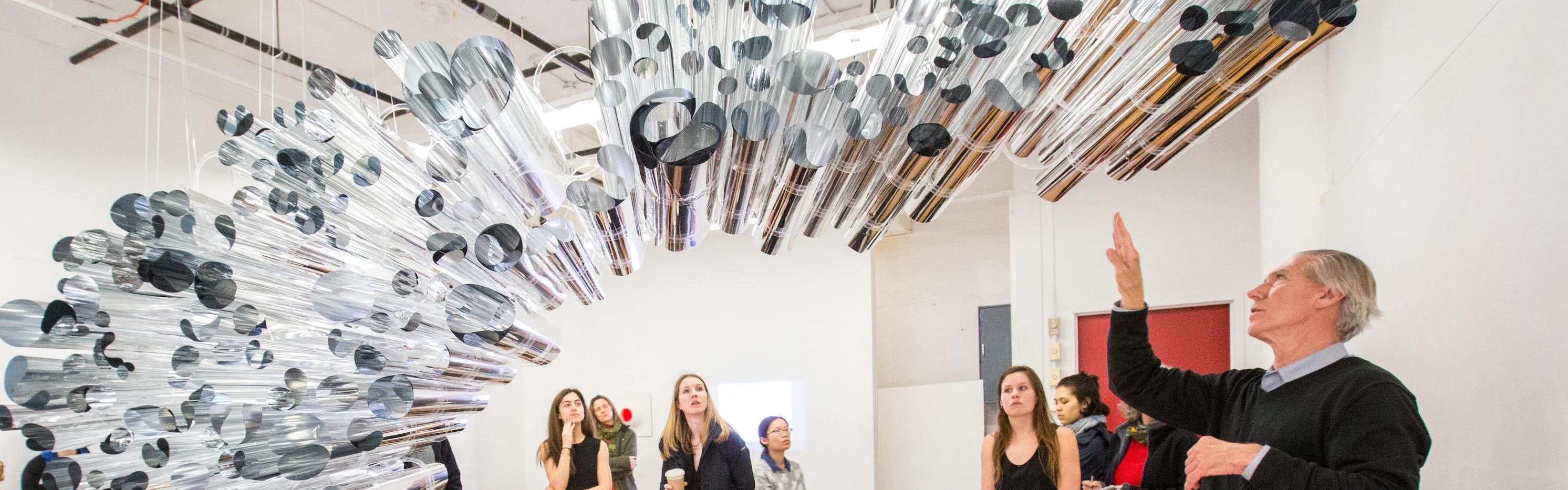 A professor explains a piece of artwork hanging in the middle of a room to a group of students