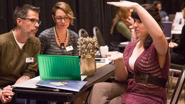 A student seated across from two adults at a table indicates a vertical dimension with both hands