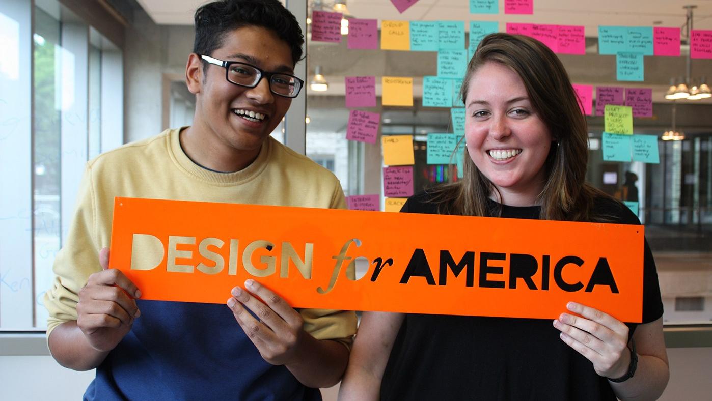 Two Design for America students hold an orange Design for America sign